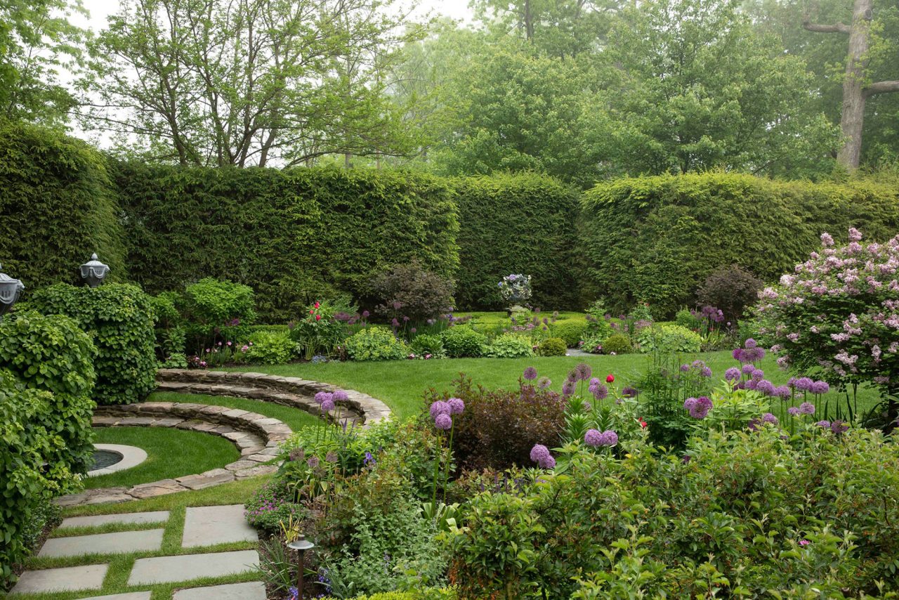 Perennial garden with fountain and lawn surrounded by tall arborvitae privacy hedge.